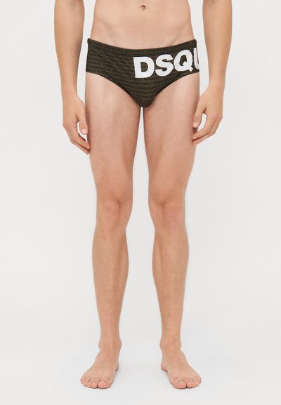 Mannelijke model in donkere slips met herhaaldelijk "DSQUARED2" tekstpatroon en grote witte "DSQ" letters aan de voorkant, staande op blote voeten.