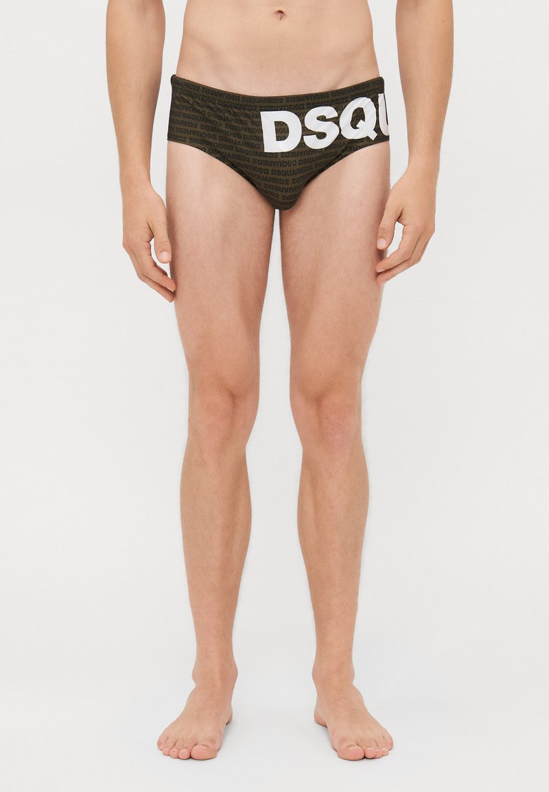 Moški model v temnih boksarkah z ponovljenim vzorcem besedila "DSQUARED2" in velikim belim napisom "DSQ" spredaj, stoji bos.