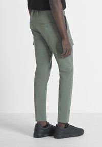 Cargo-broeken van lichtgroen materiaal met een slanke pasvorm. Voorzien van zijzakken, een knoopdetail aan de achterkant en gedragen met zwarte sneakers.
