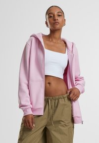 Svietiaca ružová mikina na zips s jemnou textúrou, s kapucňou a prednými vreckami, nosená otvorená cez biely crop top a khaki nohavice.