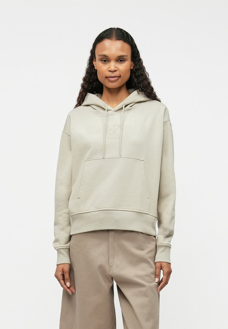 Μπεζ φούτερ με κουκούλα από μαλακό ύφασμα, που διαθέτει μπροστινή τσέπη, κορδόνια και διακριτικό λογότυπο τυπωμένο στο στήθος.