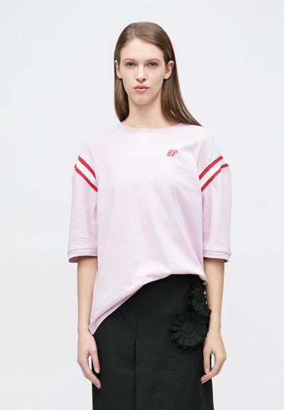 Ανοιχτό ροζ βαμβακερό t-shirt με κόκκινες ρίγες στα μανίκια και λογότυπο στο στήθος, με άνετη εφαρμογή και μανίκια γυρισμένα. Συνδυασμένο με μαύρη φούστα.