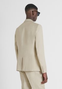 Veste de costume beige clair en tissu texturé, dotée d'une fente simple, de revers crantés et de quatre détails de boutons sur les manches.