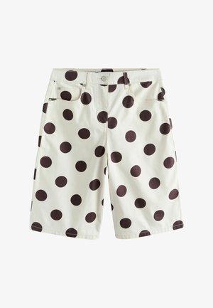 Femeie purtând ochelari de soare negri, un top cu dungi albe și negre și pantaloni scurți albi cu buline negre mari, stând în fața unui zid de beton.