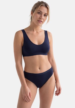 Jeune femme aux cheveux clairs portant un soutien-gorge de sport et une culotte assortis bleu foncé, posant devant un fond blanc uni.