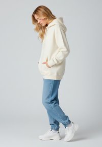 Felpa di cotone color crema con tasca a marsupio, cappuccio con coulisse e polsini a coste, abbinata a joggers in denim blu e sneakers bianche.