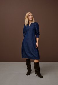 Nő áll barna háttér előtt, hosszú ujjú tengerészkék midi ruhát és sötétbarna térdig érő, magassarkú csizmát visel.