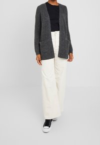Grå stickad cardigan med öppen front, ribbad textur och två framfickor, tillsammans med off-white kordfløyelsbyxor och svarta sneakers.