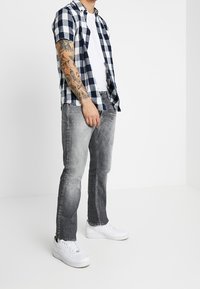Camicia a maniche corte a quadri blu e bianchi sopra una t-shirt bianca, abbinata a jeans grigi e sneaker bianchi, con tatuaggi visibili sul braccio.