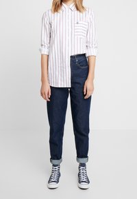 Chemise blanche à boutons avec des rayures verticales rouges, dotée d'une poche sur le côté gauche. Associée à un jean en denim bleu foncé retroussé et à des baskets montantes bleues.