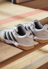 Weiße Adidas Specialist Sneaker mit schwarzen Streifen und Gummisohlen auf hölzernen Tribünen im Sonnenlicht.