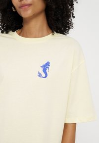 Femme portant un t-shirt jaune pâle avec un petit motif de sirène bleue sur la poitrine gauche et des boucles d'oreilles créoles dorées.