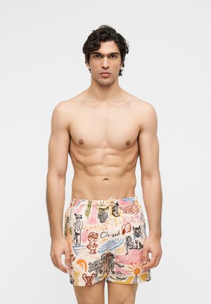 Homme torse nu aux cheveux bouclés foncés portant un short coloré avec des imprimés artistiques, debout devant un fond clair uni.