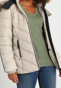 Beżowa kurtka puchowa z ukośnym wzorem pikowania, szarym kołnierzem i zamykanymi kieszeniami po bokach, noszona na zielonym swetrze dzianinowym i niebieskich dżinsach.