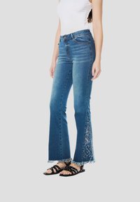 Középkék színű, bővülő szárú farmernadrág, rojtos szegélyekkel és részletes virágos hímzéssel az oldalon. Fekete pántos szandállal viselve.