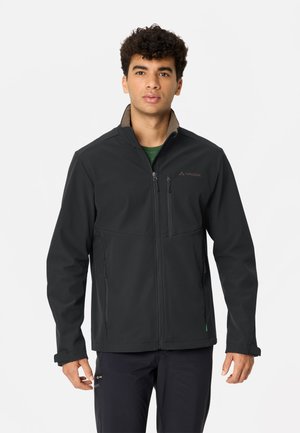 Mann mit lockigem Haar, der eine schwarze Jacke mit Reißverschluss und Brust- sowie Seitentaschen trägt, steht vor einem schlichten weißen Hintergrund.