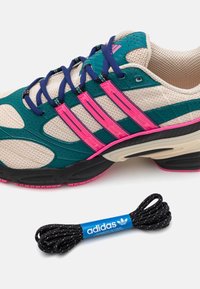 Adidas-niken har en nätöverdel i teal och beige, med rosa accenter och tre signaturstripes. Inkluderar svarta och vita skosnören.
