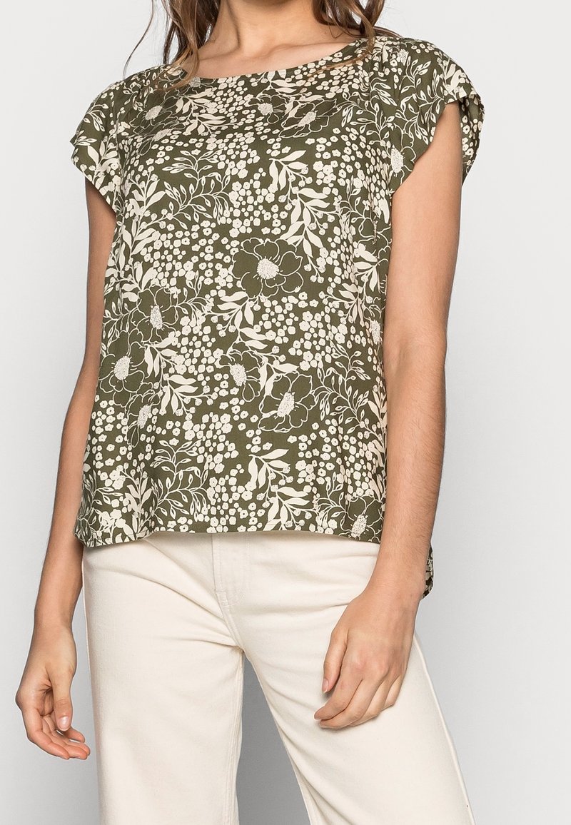 Mujer con blusa corta de manga en estampado floral verde oliva y pantalones blancos de cintura alta, de pie frente a un fondo liso.