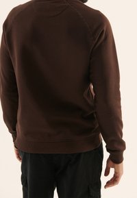 Brun sweatshirt med ribbade ärmar och fåll, med en rund halsringning och raglanärmar. Tyget verkar mjukt och texturerat.