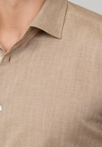 Beige herringbone patroon overhemd met een omklapbare kraag en een enkele knoopdetail. De stof lijkt licht en gestructureerd te zijn.