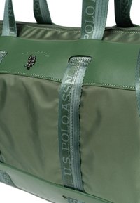 Zielona torba typu duffel wykonana z gładkiego nylonu, z subtelnym logo, kontrastującymi paskami i usztywnionym kształtem. Zamknięcie na zamek błyskawiczny u góry.