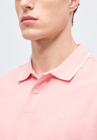 Κοντινό πλάνο ενός ατόμου που φοράει ένα ανοιχτό ροζ πόλο μπλουζάκι με γιακά, κουμπί και το λογότυπο "GANT" στο στήθος.