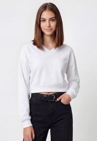Sudadera blanca recortada con escote en v, mangas largas y puños acanalados, combinada con pantalones negros de talle alto y un cinturón negro texturizado.