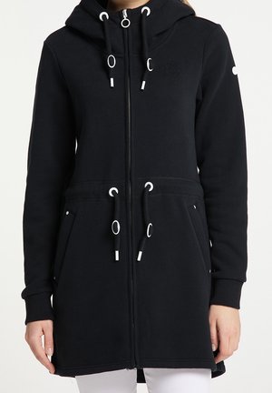 Czarna bluza z kapturem zapinana na zamek, z troczkami przy kapturze i w pasie, bocznymi kieszeniami oraz ściągaczami na mankietach, noszona na białych spodniach.