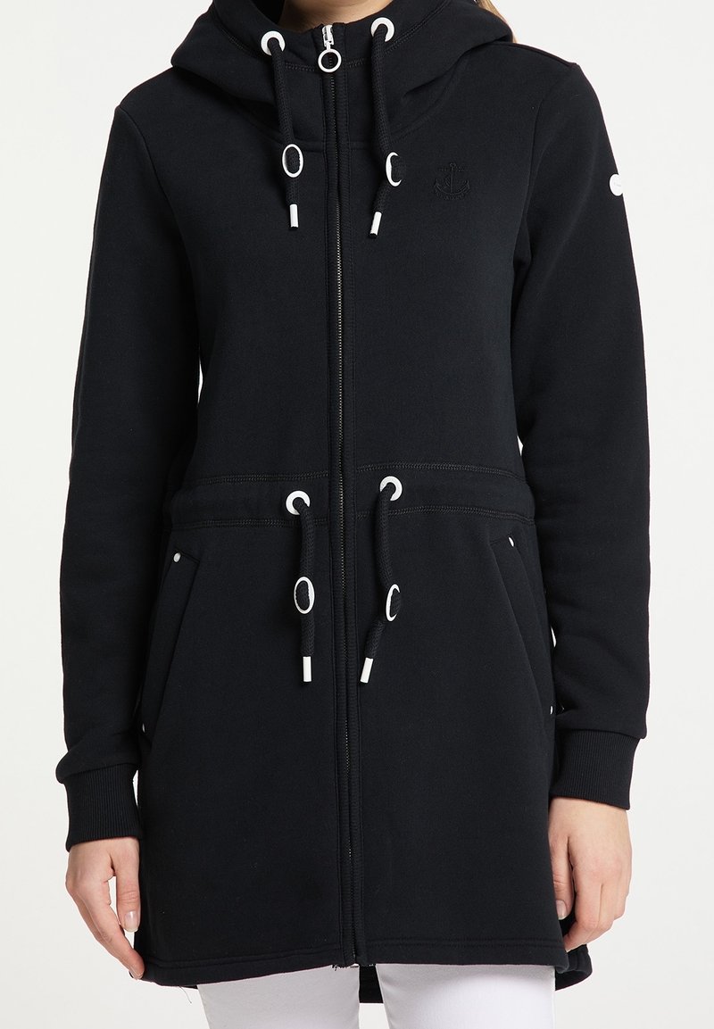 Veste zippée noire à capuche avec cordons au niveau de la capuche et de la taille, poches latérales, poignets côtelés, portée sur un pantalon blanc.