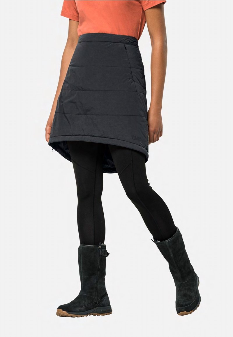 Persona con una camiseta naranja de manga corta, falda aislante negra sobre leggings negros y botas negras hasta la mitad de la pantorrilla con suelas beige.