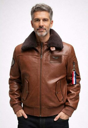 Homme aux cheveux gris et à la barbe portant une veste aviateur en cuir marron avec col en peau lainée et un porte-clés rayé sur la manche, mains dans les poches.