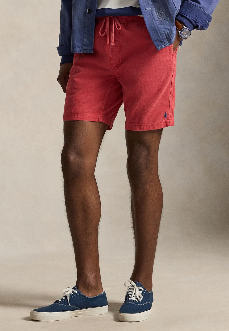 Άνδρας που φοράει κόκκινα σορτς με κορδόνια, μπλε καμβαδένια παπούτσια με λευκές σόλες, ναυτικό μπουφάν και ρολόι χειρός, στέκεται με τα χέρια στις τσέπες.