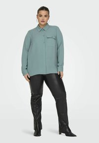Light teal button-up shirt with a collar and single chest pocket, paired with shiny black faux leather pants featuring slits at the bottom.