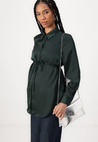 Σκουροπράσινο σατέν πουκάμισο με κουμπιά και μακριά μανίκια, δεμένο στη μέση. Ασημί μεταλλική τσάντα χειρός με αλυσίδα. Μπλε τζιν παντελόνι.