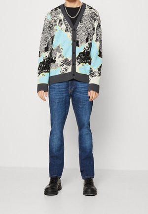 Strickjacke in Blau, Schwarz und Weiß mit Mustern, mit V-Ausschnitt, Knöpfen vorne und gerippten Bündchen, kombiniert mit dunkelblauen Jeans und schwarzen Schuhen.