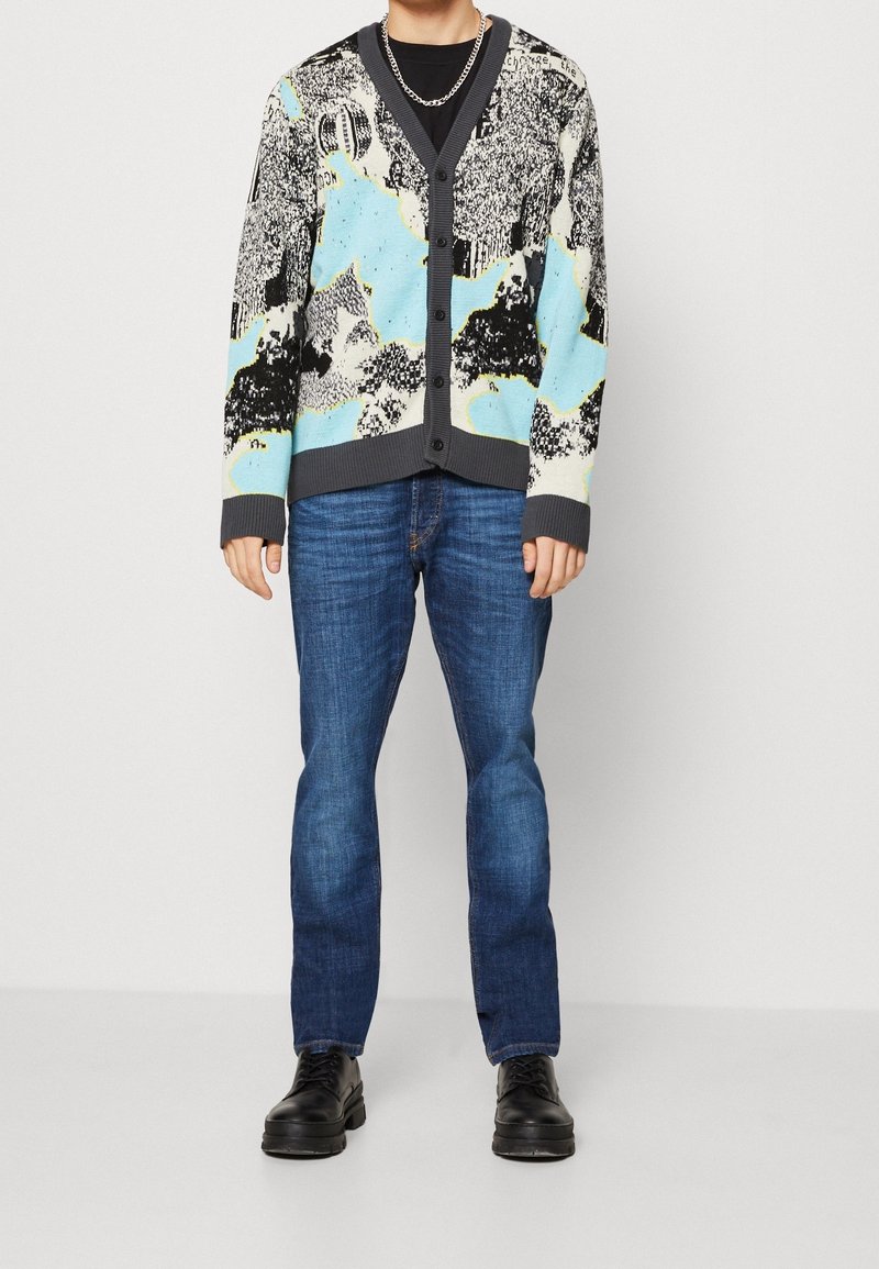 Cardigan tricoté en bleu, noir et blanc, avec un col en V, des boutons frontaux et des poignets côtelés, associé à un jean bleu foncé et des chaussures noires.