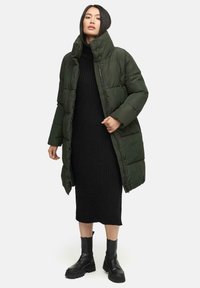 Dark green puffer coat with high collar, featuring a zip closure and quilted texture, worn over a black ribbed dress and chunky black boots.