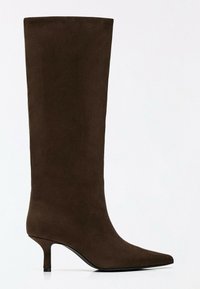 Botte haute en daim marron jusqu’au genou avec bout pointu et talon kitten fin sur fond blanc uni.
