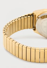 Primer plano de una pulsera de reloj de metal con tono dorado, curva sobre una superficie blanca, mostrando la parte trasera de la esfera del reloj.