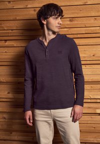 Long-sleeve purple henley shirt made of textured fabric, featuring three buttons and a small logo on the left chest, paired with light pants.