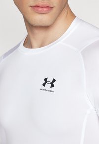 Hvid atletisk t-shirt lavet af blødt, strækbart stof. Har et sort "Under Armour" logo på brystet og syet detaljering på siderne.