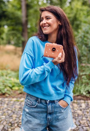 Light blue sweatshirt and distressed denim shorts. She holds a pink wallet with white polka dots and a round logo. Green trees in background.