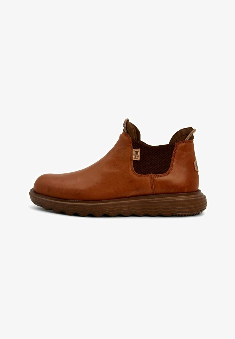 Botín de cuero marrón hasta el tobillo con paneles elásticos a los lados, suela marrón texturizada y punta redondeada. Presenta una lengüeta en la parte posterior para facilitar el uso.