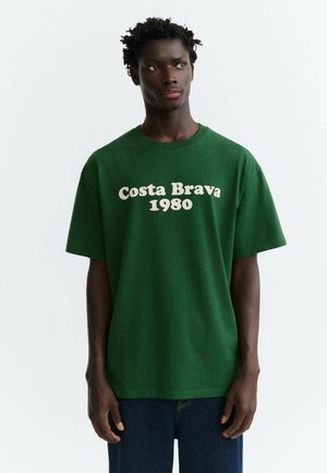 Młody mężczyzna w zielonej koszulce z napisem "Costa Brava 1980", stoi na jednolitym białym tle, patrząc na bok.