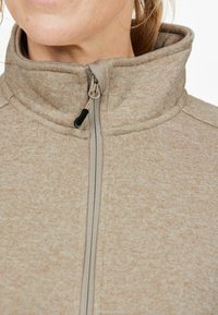 Ljusbrun fleecejacka med hög krage, som har en framzipper, texturerad yta och subtila, varierande mönster över hela.