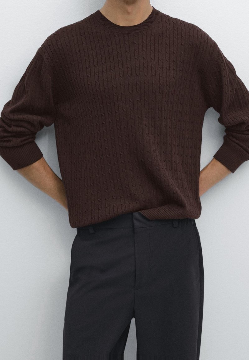Homme portant un pull torsadé marron foncé à manches longues et un pantalon noir, debout, les mains derrière le dos.