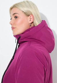 Blonde woman in profile wearing silver hoop earrings and a bright purple hooded jacket with black drawstrings against a plain white background.