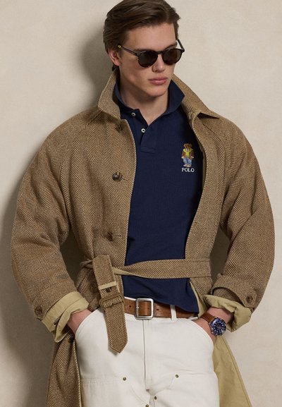 Trench-coat en chevron marron avec une ceinture nouée à la taille, polo bleu marine avec un logo d'ours brodé, pantalon crème, et lunettes de soleil foncées. Montre en cuir marron.