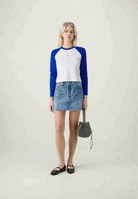 White long-sleeve shirt with blue sleeves, paired with a denim mini skirt. Black bag with a patterned design and black flat shoes.