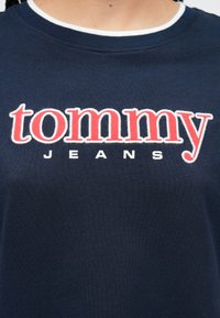 Lähipilt tumesinisest särgist, mille rinnal on suured punased tikitud tähed "tommy" ja väiksemad valged "JEANS" tähed.